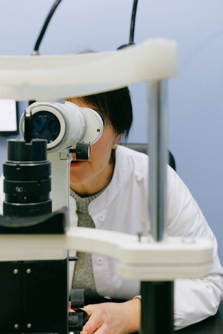 Anonymous female doctor in white coat observing object through big microscope while sitting in light doctor office with blue walls