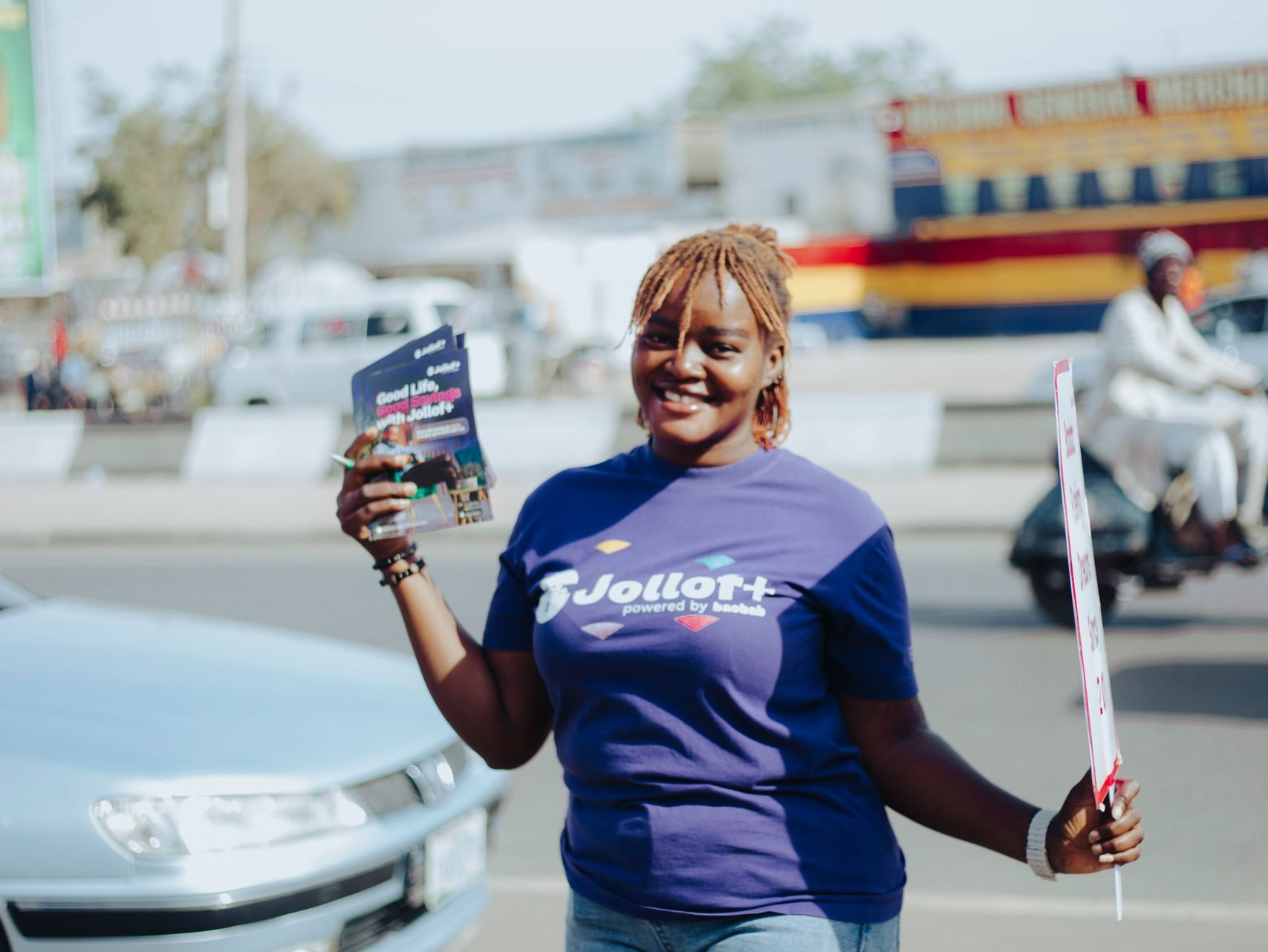 Cheerful woman wearing a purple T-shirt and jeans, holding flyers on a sunny day.