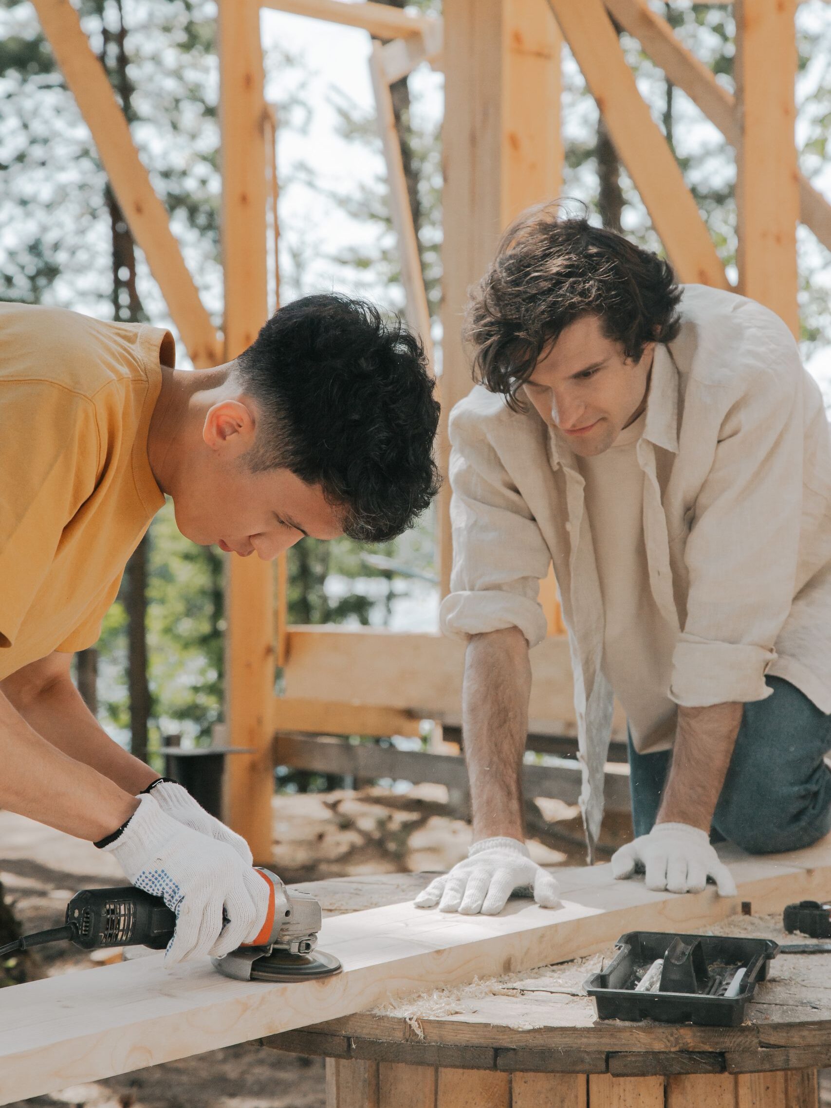 Two men collaborate to sand and polish a wooden beam at a construction site outdoors.